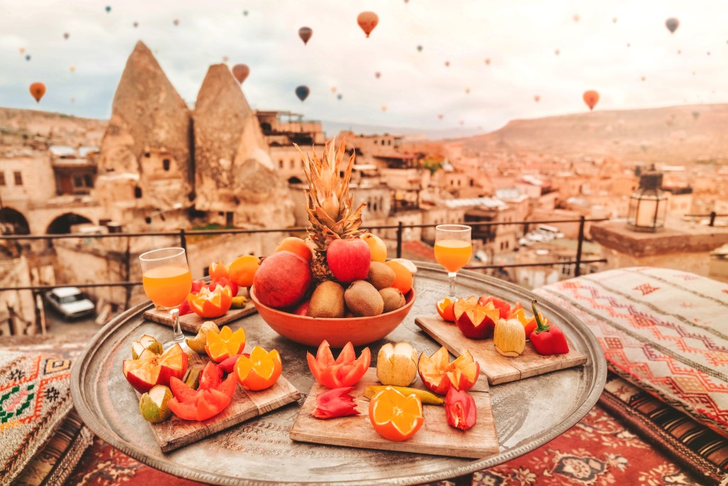 Bir tepsi üzerinde çeşitli meyveler ve meyve suyu, arka planda sıcak hava balonlarıyla Kapadokya manzarası.